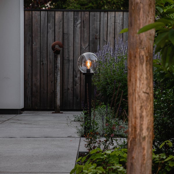 Moderne Wegeleuchte mit schwarzem Mast und transparenter Glaskugel auf einer Terrasse mit Steinplatten vor einem Holzzaun.