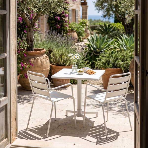 Mediterrane Terrasse mit weißem quadratischem Bistrotisch und zwei weißen Gartenstühlen aus Metall zwischen Terrakottakübeln und Pflanzen.
