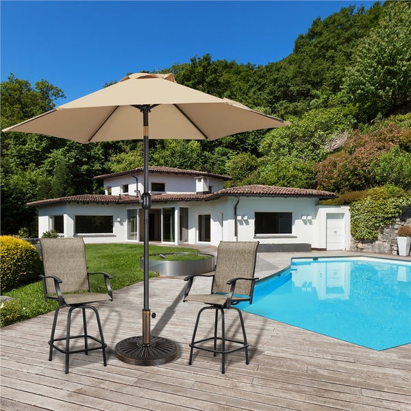 Großer beiger Sonnenschirm mit zwei passenden Barstühlen auf einer Holzterrasse neben einem Pool in einem hellen, modernen Garten.