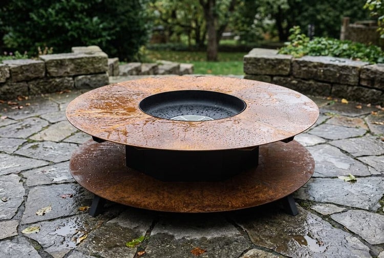 Runde Feuerstelle mit Grillplatte aus Metall in Rostoptik auf einer Steinterrasse im Garten