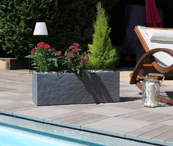 Rechteckiger Pflanzkasten in Steinoptik mit roten Blumen und kleiner Konifere auf einer Terrasse am Pool.