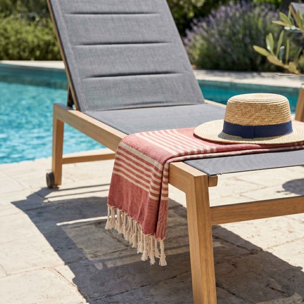 Gartenliege aus Holz mit grauer Liegefläche an einem Pool, dekoriert mit einem gestreiften Tuch und einem Strohhut.