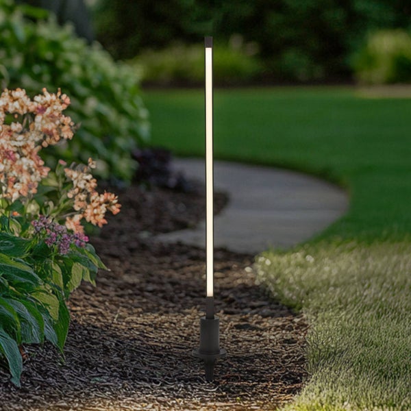Moderne, schlanke LED-Gartenleuchte mit Erdspieß in einem Gartenbeet mit Mulch neben einem Rasen und einem Gartenweg.