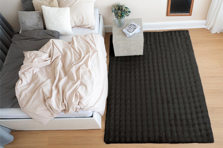 Ein rechteckiger Teppich mit strukturierter Oberfläche liegt neben einem Bett in einem modern eingerichteten Schlafzimmer mit Parkettboden.