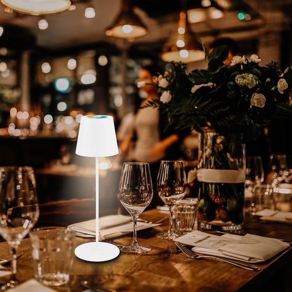 Tischszene mit einer weißen Tischlampe, Gläsern, Besteck und Blumenvase.