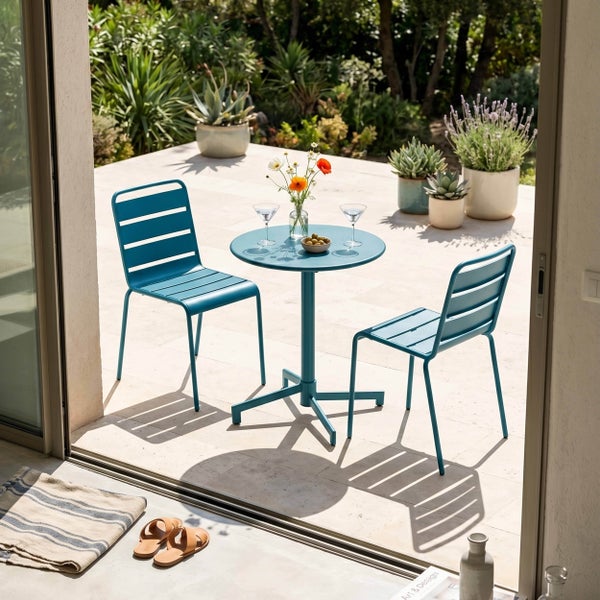 Set aus rundem Metall-Bistrotisch und zwei Metallstühlen mit Latten-Design auf einer hellen Terrasse mit Topfpflanzen im Hintergrund.