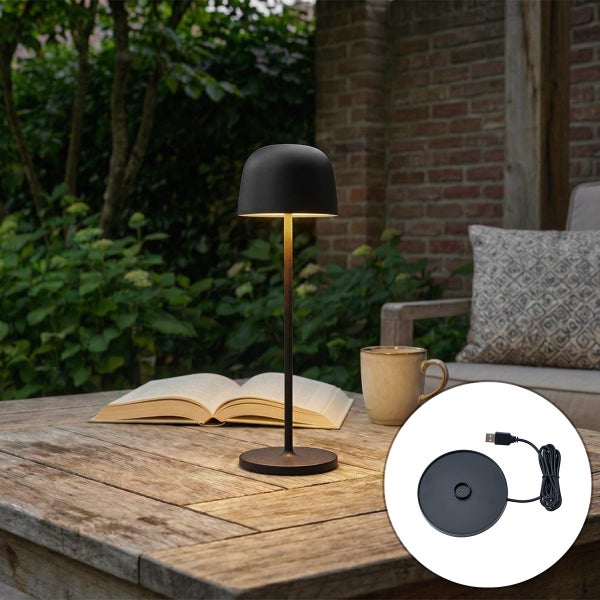 Schwarze, kabellose LED-Tischleuchte auf einem Holztisch im Außenbereich mit Buch und Tasse, inklusive USB-Ladestation.