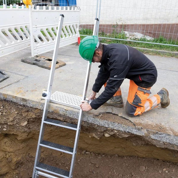 Ein Bauarbeiter montiert eine Grabenleiter aus Aluminium mit Podest und Handlauf am Rand einer Baugrube.
