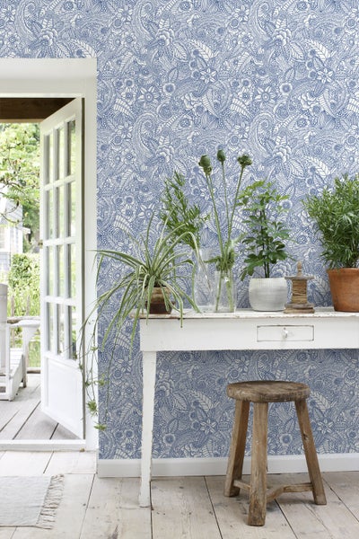 Zimmer mit blau gemusterter Tapete, weißem Tisch und Holzschemel