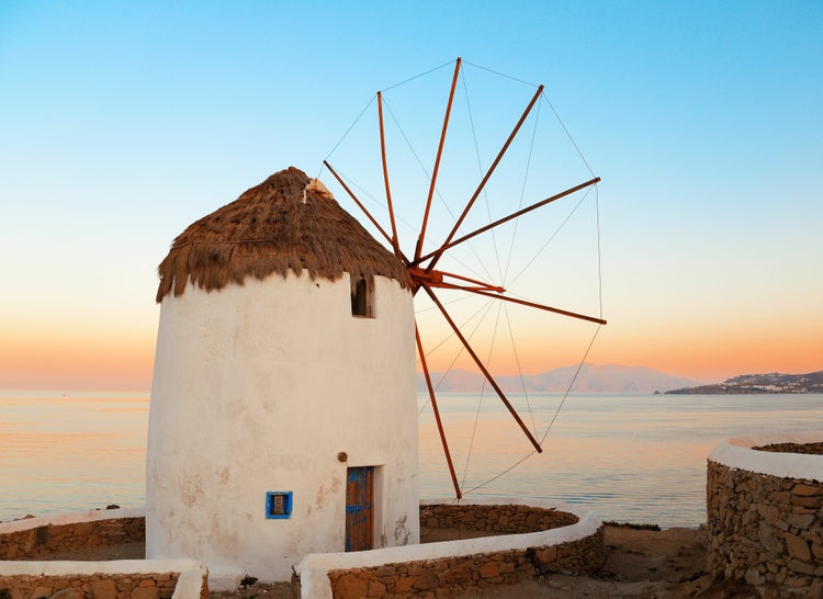 Griechische Windmühle mit weißer Fassade, Reetdach und Holzflügeln an einer Steinküste vor dem Meer bei Sonnenuntergang.