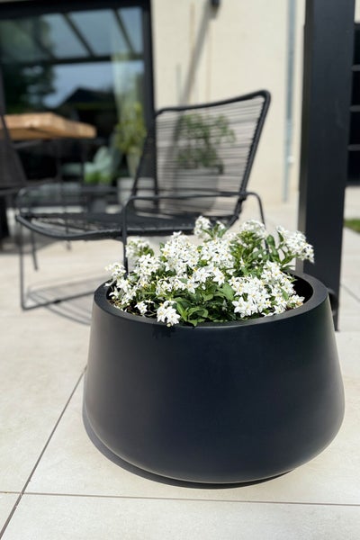 Schwarzer runder Pflanzkübel mit weißen Blumen auf einer hellen modernen Terrasse neben einem schwarzen Lounge-Sessel.