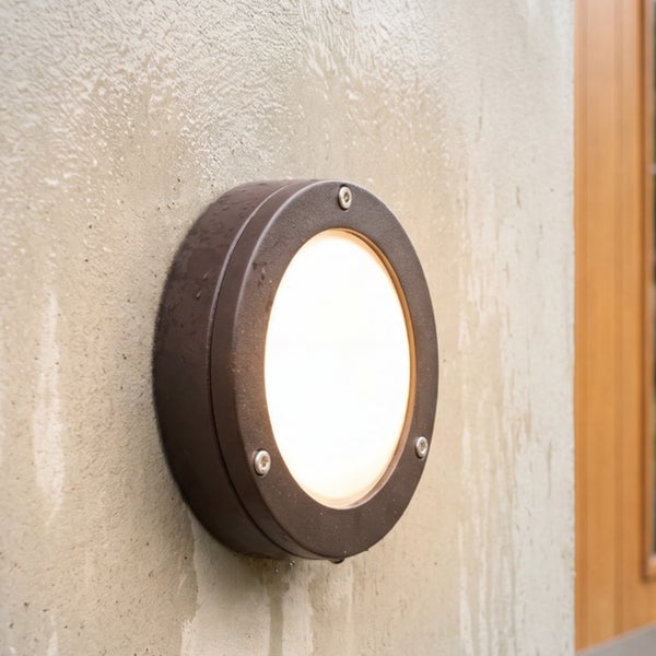 Runde Außenwandleuchte aus dunkelbraunem Metall mit warmweißem Licht an einer Hauswand.