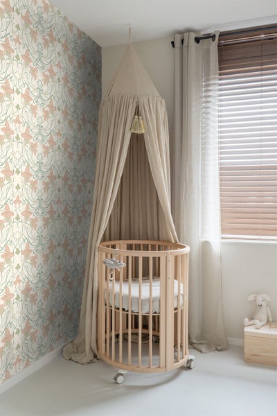 Helles Kinderzimmer mit rundem Babybett aus Holz, Stoffhimmel und einer Wand mit floraler Mustertapete sowie Fenster mit Jalousien.