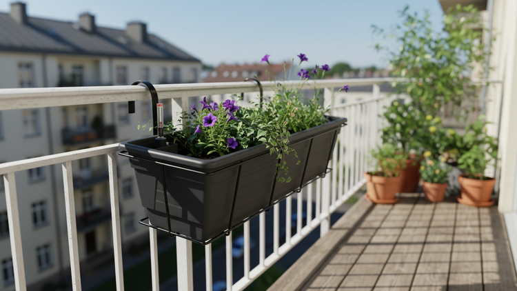 Schwarzer Balkonkasten aus Kunststoff mit Bewässerungssystem und violetten Blumen an einem weißen Geländer auf einem hellen Balkon.