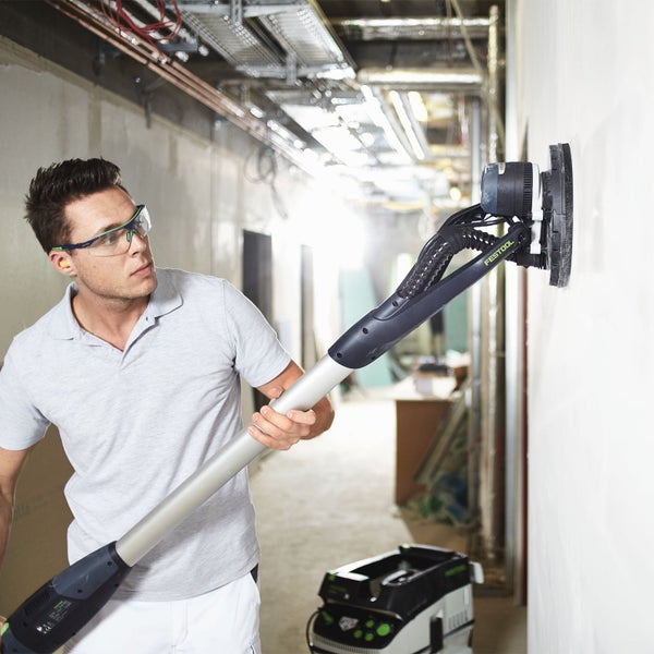 Ein Mann schleift eine Wand mit einem Langhalsschleifer von Festool, während ein Staubsauger daneben steht.
