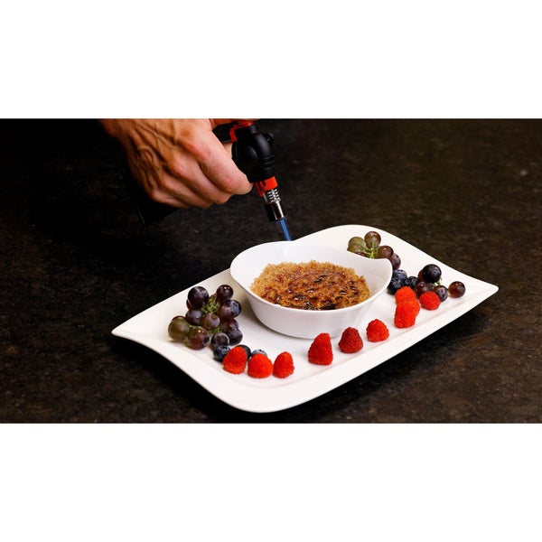 Ein Mann karamellisiert eine Crème brûlée mit einem Gasbrenner, serviert auf einem Teller mit Trauben, Blaubeeren und Himbeeren.