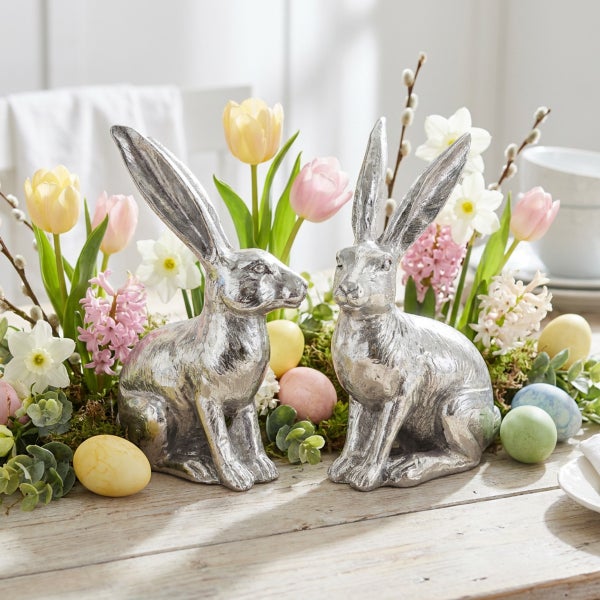 Zwei Osterhasen Figuren aus Metall als Dekoration mit Frühlingsblumen und Ostereiern auf einem Holztisch für die festliche Tischdekoration.
