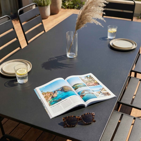 Moderner Gartentisch mit passenden Metallstühlen auf einer Holzterrasse, dekoriert mit Magazin, Gläsern und einer Vase.