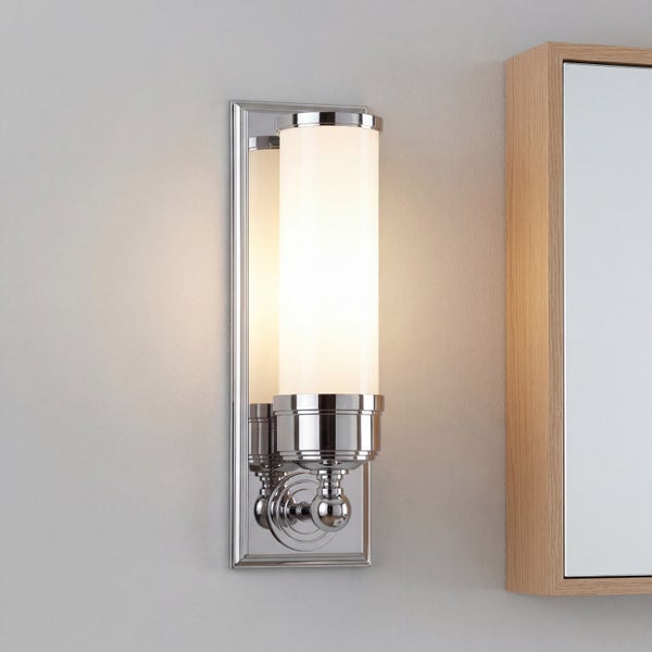 Moderne Wandleuchte im Badezimmer mit glänzendem Chrom-Sockel, zylindrischem Opalglas und warmem Licht neben einem Spiegel.
