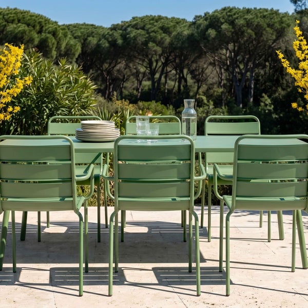 Modernes Gartenmöbel-Set aus grünem Metall mit Tisch und Stühlen auf einer hellen Steinterrasse vor einer Kulisse aus Bäumen.