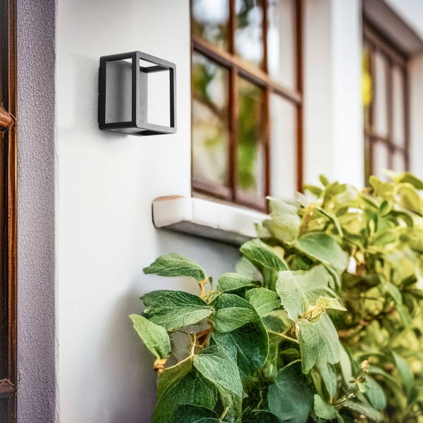 Moderne schwarze würfelförmige Außenwandleuchte an einer weißen Hauswand neben einem Fenster und grünen Pflanzen.