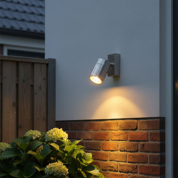 Moderne Außenwandleuchte aus Edelstahl als Strahler, beleuchtet eine Ziegelmauer im Außenbereich neben einem Holzzaun und Hortensien.