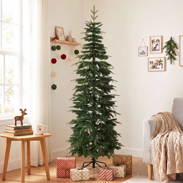 Ein hoher künstlicher Weihnachtsbaum steht in einem wohnlich eingerichteten Raum mit Geschenken darunter neben einem Sofa und einem Beistelltisch.