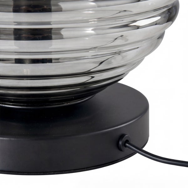 Nahaufnahme einer Tischleuchte mit mattschwarzem Standfuß, schwarzem Netzkabel und einem horizontal gerippten Glasschirm.