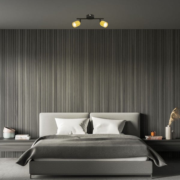 Modernes Schlafzimmer mit grauem Polsterbett, dunkler Lamellenwand und schwarzem Deckenstrahler mit zwei goldfarbenen Leuchtenköpfen.