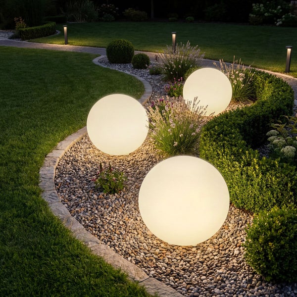 Drei warmweiß leuchtende Kugelleuchten in einem Gartenbeet mit Kies, Pflanzen und Rasen bei Nacht.