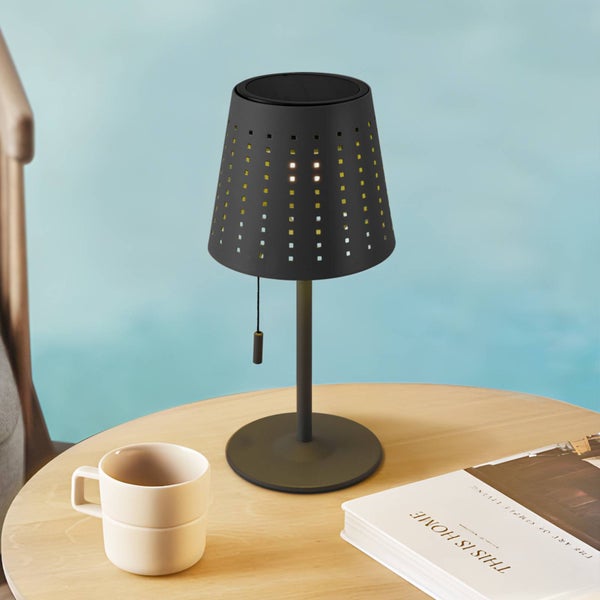 Solar-Tischleuchte mit perforiertem Schirm, rundem Standfuß und Zugschalter auf einem Holztisch neben einer Tasse und einem Buch.