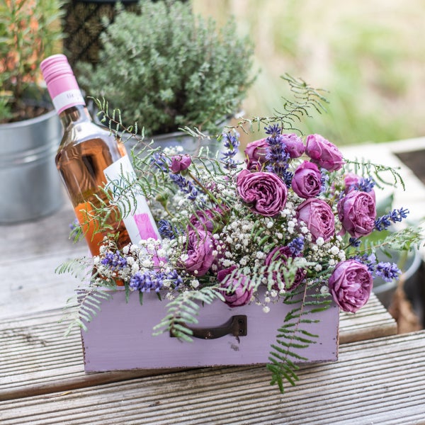 Dekorative Holzkiste mit Rosen, Lavendel, Schleierkraut und einer Flasche Roséwein.