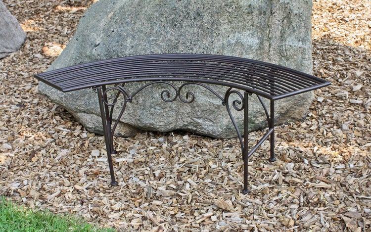 Gebogene Gartenbank aus Metall mit Latten-Sitzfläche und dekorativen Verzierungen vor einem Stein auf Rindenmulch.