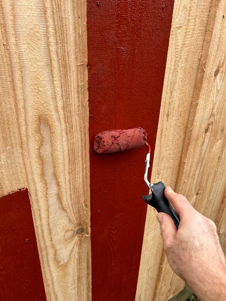 Hand streicht Holzlatten mit einer Farbwalze und roter Holzfarbe.