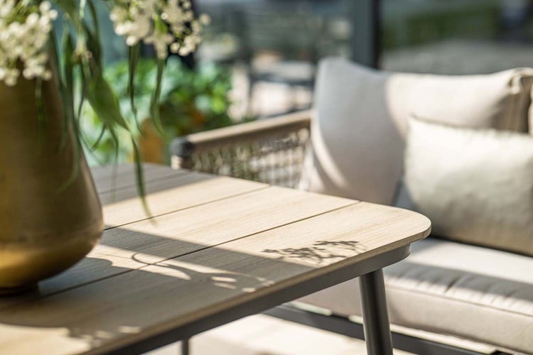 Moderner Gartentisch mit Tischplatte in Holzoptik und Metallgestell auf einer hellen Terrasse neben einem Gartensofa mit Kissen.