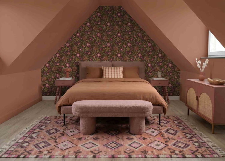 Gemütliches Schlafzimmer mit Dachschrägen, floraler Tapete, einem Bett in Terrakotta, gemustertem Teppich und einer gepolsterten Bank.
