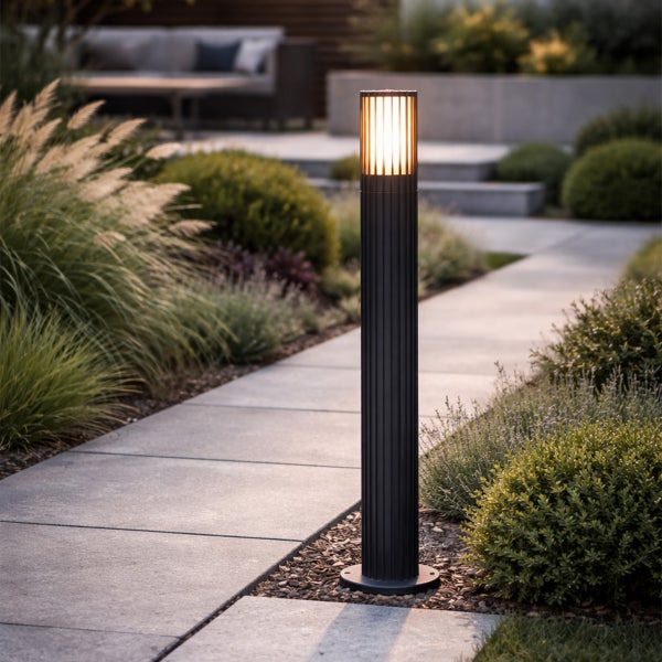 Schwarze Pollerleuchte mit geriffeltem Design und warmem Licht an einem Gartenweg aus Steinplatten zwischen Gräsern und Sträuchern.