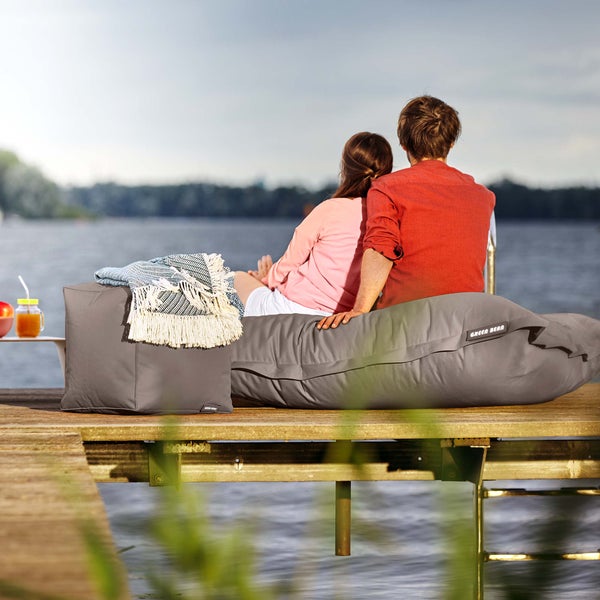 Ein Paar sitzt auf einem grauen Sitzsack und einem Sitzwürfel von Green Bean auf einem Holzsteg am Wasser.