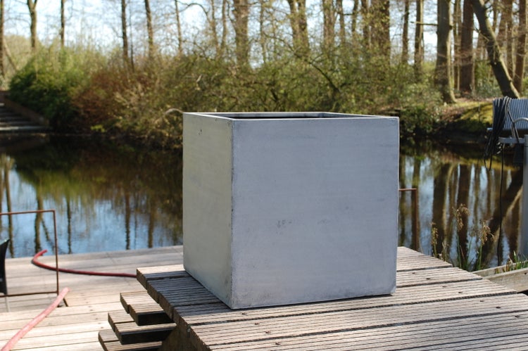 Quadratischer Pflanzkübel in Beton-Optik auf einem Holzsteg an einem Gewässer.