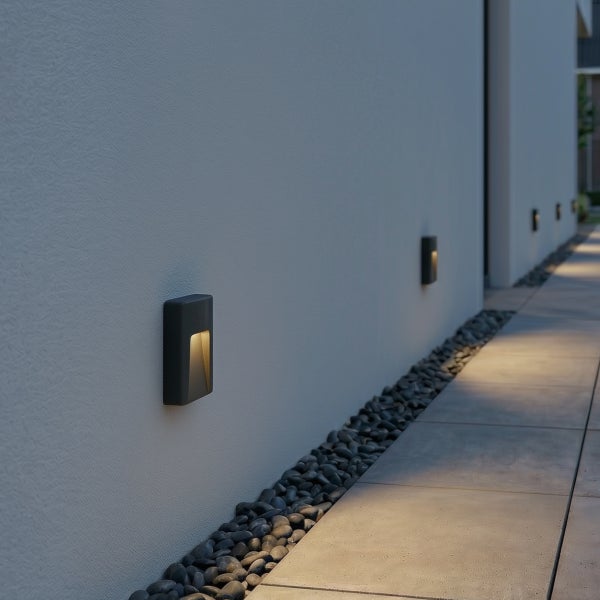 Moderne rechteckige LED Wandleuchten für den Außenbereich an einer Fassade, die einen Gehweg mit Kieselsteinen und Steinplatten beleuchten.