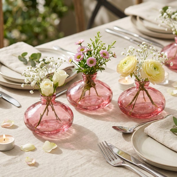 Drei kleine bauchige Glasvasen als Tischdekoration bestückt mit frischen Blumen auf einem gedeckten Tisch.