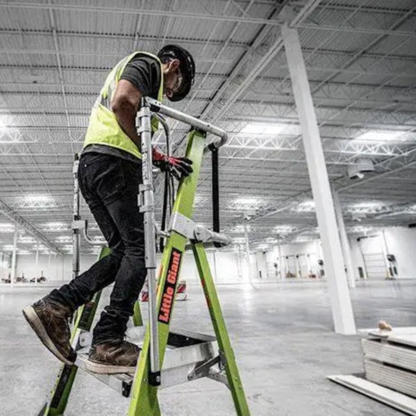 Handwerker mit Schutzhelm und Warnweste steht auf einer Little Giant Stehleiter in einem großen Lagerhaus.