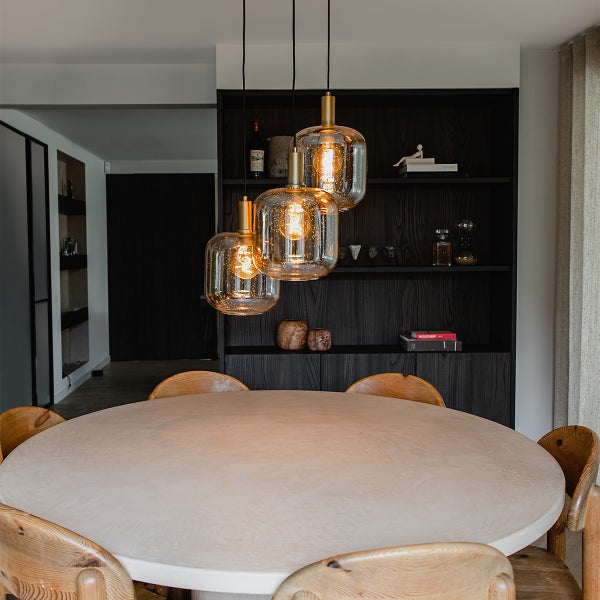 Szene mit drei Pendelleuchten über einem Esstisch und einem dunklen Regal im Hintergrund.