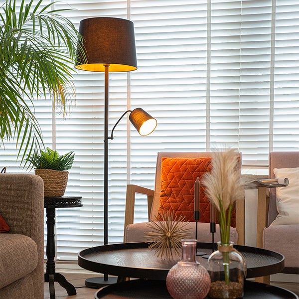 Moderne Stehleuchte mit schwarzem Textilschirm und Lesearm in hellem Wohnzimmer mit Jalousien, Sesseln und Beistelltischen.
