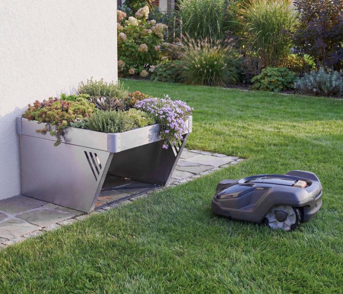 Mähroboter-Garage aus Metall mit integriertem Pflanzdach voller Sukkulenten und Blumen auf einem Rasen neben einem Mähroboter im Garten.