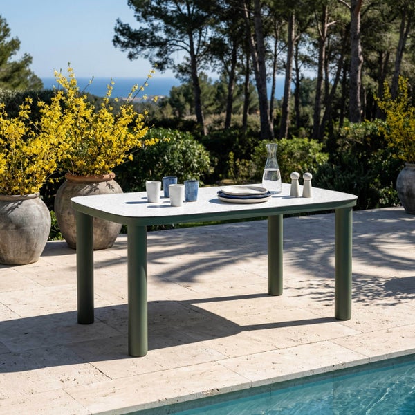 Moderner Gartentisch mit grünen Metallbeinen auf einer hellen Fliesenterrasse neben einem Pool, umgeben von gelben Blumen und Blick auf das Meer.