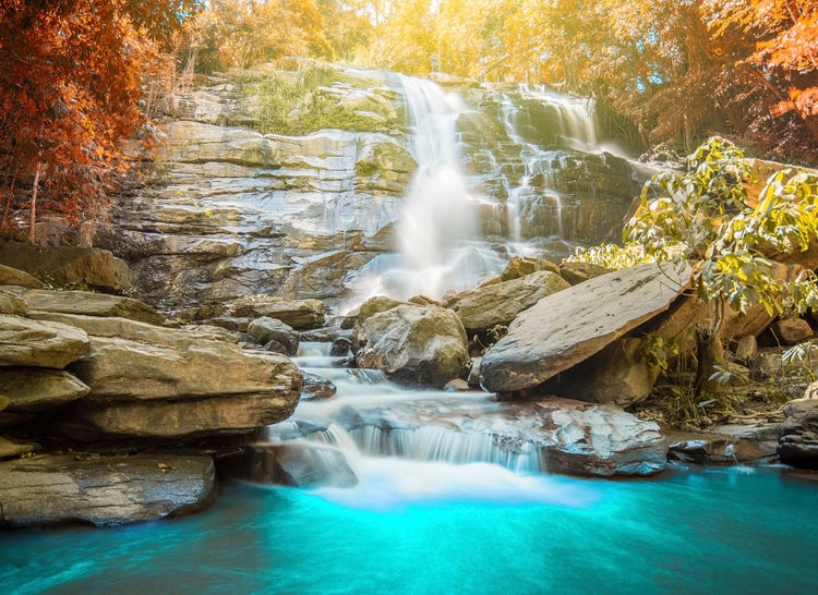 Fototapete mit Wasserfall über Felsen in blaues Wasser, umgeben von herbstlichem Wald.