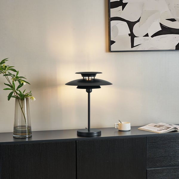 Moderne schwarze Metall-Tischleuchte mit mehrlagigem Schirm auf einem dunklen Sideboard neben einer Vase, Tasse und Zeitschrift.