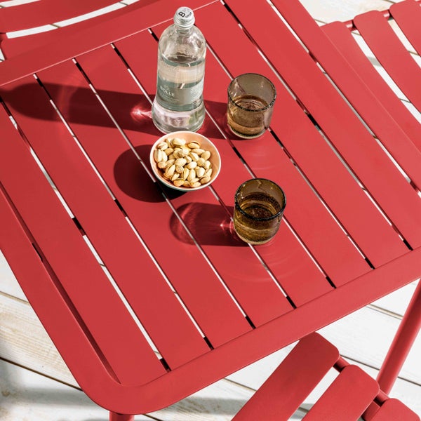 Roter quadratischer Metall-Gartentisch in Latten-Optik mit einer Wasserflasche, zwei Gläsern und einer Schale Pistazien auf einer Holzterrasse.
