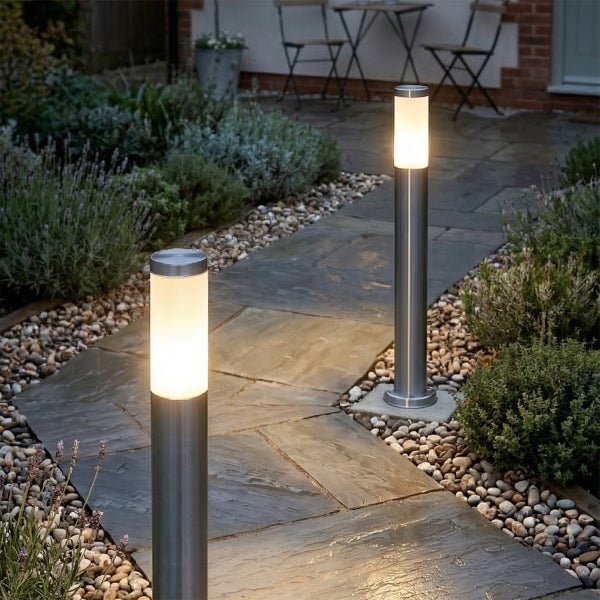 Zwei moderne Wegeleuchten aus Edelstahl mit warmweißem Licht beleuchten einen Gartenpfad aus Steinplatten und Kies.